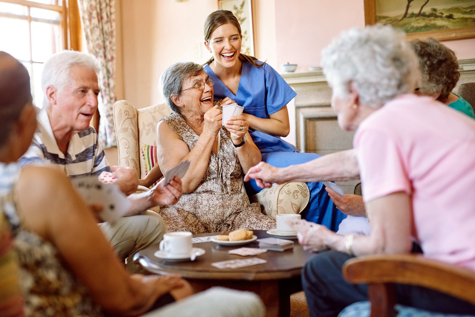 Seniori hrajú karty v zariadení a ošetrovateľka na nich dohliada